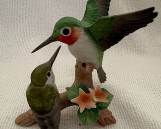 This vintage ceramic figurine depicts two hummingbirds perched on a branch adorned with small, star-shaped flowers. The larger hummingbird, positioned above and to the right, is shown with wings outstretched. It features vibrant green plumage on its head and back, a white chest, and a striking reddish-orange throat patch. Its eye is a distinct black dot. The smaller hummingbird, to the left and below, is depicted with its head tilted upwards, seemingly interacting with the larger bird. It is primarily a muted green color with a white chest and a black eye. The branch is rendered in a textured brown, and the flowers are a soft peachy-orange with green leaves. The figurine is marked as 'Andrea by Sadek' and 'Japan'.