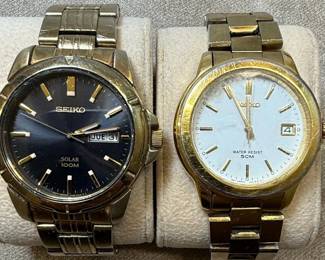 This lot features two men's Seiko wristwatches. The first is a solar-powered watch with a dark blue dial, gold-tone hour markers and hands, and a day-date complication. It indicates 'SOLAR 100M' on the dial and 'MOV'T JAPAN V158-0AD0 R 2' on the case back, which is stainless steel. The second watch has a white dial with gold-tone hour markers and hands, and a day-date complication, and 'WATER RESIST 50M' printed on the dial. Its case back indicates 'MOV'T JAPAN V742-2161 R 2'. Both watches have gold-tone metal bracelets and cases. The overall aesthetic suggests a classic, functional design typical of Seiko's offerings in the late 20th or early 21st century. The crystals on both watches show significant scratching.