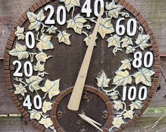 This decorative garden thermometer and clock features a circular design with a rich, dark brown base and raised accents of green grape leaves and vines, consistent with the Fossil Sumac motif. The outer rim is textured to resemble brickwork, and the numbers indicating temperature are large and white, ranging from 0 to 100 degrees Fahrenheit. A smaller dial at the bottom displays hours from 1 to 12, functioning as a clock. The thermometer's needle is a pale, weathered gold color, and it appears to be made of a durable material suitable for outdoor use, such as cast resin or metal, designed to withstand the elements. Based on similar listings, this item is likely from Whitehall Products and features their 'Fossil Sumac' design.