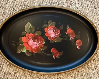This is an oval-shaped decorative tray, likely made of metal with a black enameled finish. It features a hand-painted floral design of pink and red roses with green leaves and stems, rendered in a somewhat stylized, folk-art manner. A thin gold-toned rim encircles the tray's edge. The underside of the tray bears a "Fine Arts Studio, HAND PAINTED, PHILAD'A, PA." sticker, indicating its origin and decorative nature. The overall aesthetic suggests a mid-20th century decorative piece, possibly used for serving or display.