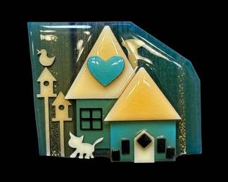 This charming brooch features a whimsical house scene crafted from layered resin and other materials. The design depicts a stylized house with a turquoise heart above the roof, a small white cat, and two birdhouses with a bird perched on one. The background is a gradient of deep blues and greens, with flecks of gold glitter adding a touch of sparkle. The overall aesthetic is folk art inspired, with a playful and colorful presentation. The back of the brooch reveals a metal pin backing and a label identifying the maker as 'HOUSE PINS® by Lucinda' with contact information.