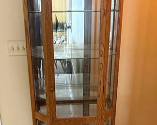 This is a traditional corner curio cabinet constructed from oak wood, featuring a multi-sided glass display area. The cabinet has a stepped top with decorative molding and a mirrored back panel, visible through the glass. It includes several adjustable glass shelves for displaying collectibles. A single interior light fixture is present at the top of the cabinet. The overall style suggests a late 20th-century or early 21st-century design, likely intended for residential use to showcase decorative items. The wood appears to be solid oak or oak veneer, with a medium-tone stain.