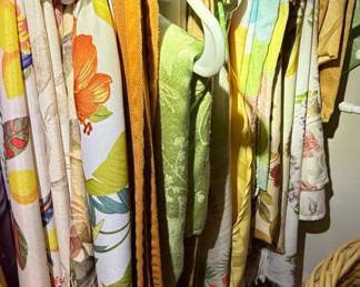 This collection features a variety of vintage fabric items, likely scarves or lengths of fabric, displayed hanging on hangers in a closet. The textiles showcase a range of vibrant floral and abstract patterns, with prominent colors including yellow, orange, green, purple, and pink. Materials appear to be lightweight, possibly silk, rayon, or cotton blends, judging by the drape and texture visible. Some pieces exhibit a textured weave, while others have smooth, printed surfaces. The overall aesthetic suggests a bohemian or mid-century modern influence, with bold graphics and nature-inspired motifs.
