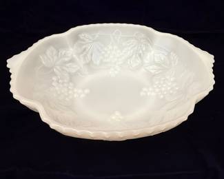 This is a white milk glass bowl featuring an embossed grape and leaf pattern, commonly known as the "Grape Harvest" pattern. It has a scalloped rim and two small handles. This pattern is a popular design by Anchor Hocking, a well-known manufacturer of milk glass.
