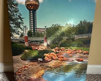 This canvas print depicts a scenic view of World's Fair Park in Knoxville, Tennessee, prominently featuring the iconic Sunsphere. The image captures a tranquil water feature with rocks and greenery in the foreground, leading the eye towards the distinctive golden sphere atop its towering structure. The sky above is a clear blue with scattered clouds, and lush trees frame the scene. The print is likely intended to evoke a sense of place and memory for those familiar with the landmark, which was originally built for the 1982 World's Fair.
