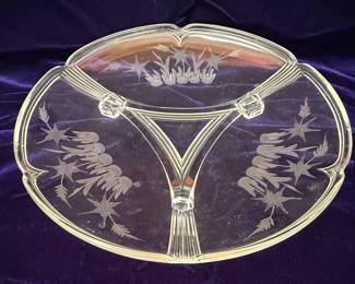 This vintage serving tray is crafted from clear glass and features a distinctive Art Deco design. It is divided into three sections by raised glass dividers that converge at the base. The rim of the tray is scalloped, and each section is adorned with etched frosted glass decoration depicting stylized floral or botanical motifs, possibly bamboo or reeds, with star-like elements. The overall aesthetic suggests a piece from the Art Deco period, likely dating from the 1920s to 1940s, intended for serving appetizers, relishes, or small desserts. The glass exhibits a slight amber or yellow tint, common in glassware of this era.