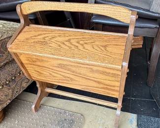 This is a vintage "Priscilla" style sewing box stand crafted from solid oak, featuring a warm, natural finish that highlights the wood's grain. The design is functional and classic, with a hinged lid that conceals storage space for sewing supplies, and an open section below that could hold magazines or other items. The piece has a sturdy construction with visible joinery and a slightly curved handle/top rail. Its overall aesthetic suggests a mid-20th century origin, possibly in a farmhouse or traditional style. The wood exhibits a typical oak grain pattern with variations in color and texture. The "Priscilla" style is characterized by its standing design and often includes a lift-top compartment and an open lower shelf or magazine rack.