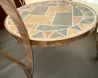 This accent table features a round, tempered glass top that sits above a decorative mosaic inlay on a lower shelf. The mosaic features geometric patterns in earthy tones of brown, beige, gray, and terracotta, bordered by a darker grout. The table's base is constructed from metal with a distressed, bronzed finish, featuring elegantly curved legs that support the structure. The combination of glass, mosaic tile, and metal creates a piece that blends modern and rustic aesthetics, suitable for various interior design styles.