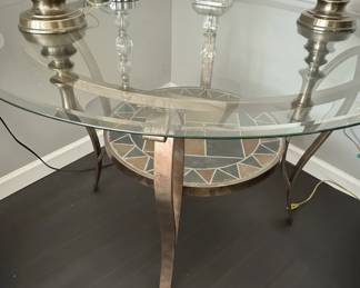 This accent table features a round, tempered glass top that sits above a decorative mosaic inlay on a lower shelf. The mosaic features geometric patterns in earthy tones of brown, beige, gray, and terracotta, bordered by a darker grout. The table's base is constructed from metal with a distressed, bronzed finish, featuring elegantly curved legs that support the structure. The combination of glass, mosaic tile, and metal creates a piece that blends modern and rustic aesthetics, suitable for various interior design styles.