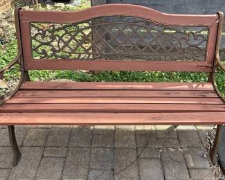 This garden bench features a decorative cast iron frame with scrolled armrests and a central back panel adorned with an intricate, lattice-like pattern. The seat and backrest are constructed from wooden slats, painted a muted reddish-brown. The cast iron elements exhibit a weathered, aged patina, suggesting outdoor use. The overall design evokes a Victorian or early 20th-century aesthetic, suitable for garden or patio seating. This specific bench appears to be the 'Tea Rose' model by Oakland Living.