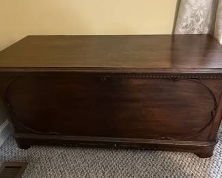 This is a vintage cedar-lined blanket chest, likely dating from the mid-20th century, manufactured by the Lane Company. The exterior is constructed from dark stained wood, possibly walnut, featuring a curved lid and decorative carved or applied molding around the base and an oval motif on the front panel. The interior is lined with aromatic cedar wood, a traditional material for protecting textiles from moths and moisture. The chest has a lock mechanism and is supported by short, carved feet. Its design suggests a piece of functional furniture that also served as a repository for personal belongings. Many Lane cedar chests from this era feature a "Style" number and manufacturing date stamped inside, which can help to further refine the identification.