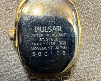 Pulsar and Caravelle Womens WristwatcThis lot features two women's wristwatches: a gold-tone Pulsar with a mother-of-pearl dial and a silver-tone Caravelle with a white dial. The Pulsar watch has an oval case and a multi-link bracelet, with 'PULSAR', 'WATER RESISTANT', 'ST. STEEL', '1N00-X139 R2', 'MOVEMENT JAPAN', and '500106' visible on the case back. The Caravelle watch has a round case, Roman numerals, and a metal expansion band, with 'CARAVELLE', 'WATER RESISTANT', and 'T SWISS T' visible on the dial. Both appear to be quartz-powered and show signs of wear consistent with age and use.hes third image