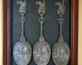 This framed set features three decorative pewter spoons, each embossed with scenes attributed to the Dutch Golden Age painter Jan Steen. The spoons are mounted on a dark backing within a wooden frame. Each spoon bowl depicts a lively scene with figures, characteristic of Jan Steen's genre paintings, and the word 'HOLLAND' is visible on the rim. The handles are adorned with figural finials, possibly birds or mythical creatures, connected by intricate scrollwork. The scenes appear to depict social gatherings or domestic life, common themes in Jan Steen's work. The spoons exhibit a dark, aged patina consistent with pewter.