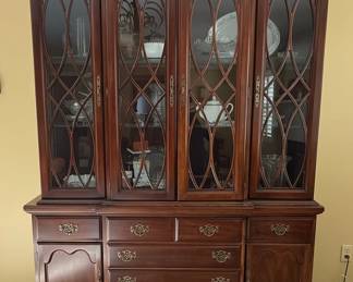 This is a large, traditional china cabinet manufactured by Schwanek Furniture, a company known for its "Fine Interiors" line. The cabinet is constructed from dark-stained wood, likely mahogany or cherry, with a rich, warm finish. It features a multi-paneled glass upper section with decorative mullions forming an intricate diamond pattern, designed to display china and decorative items. The lower section comprises several drawers and cabinets, offering ample storage. The hardware appears to be ornate brass, consistent with traditional furniture styles. The overall aesthetic suggests a piece from the mid to late 20th century, designed to be a focal point in a dining room or living area.