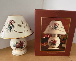This is a Lenox "Winter Greetings" candle lamp, designed to hold a tea light or votive candle. The lamp features a porcelain base and shade, both adorned with a festive winter motif. The design includes a cardinal, holly, pine sprigs, and a red ribbon, rendered in a delicate hand-painted style. The edges of the shade and the base are accented with a gold trim. The shade has cut-out details that would allow light to filter through, creating a warm ambiance. The item comes with its original box, which displays the "Winter Greetings Candle Lamp" title and a depiction of the lamp. The specific design prominently features a red cardinal.
