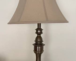 This is a Stiffel traditional table lamp featuring a metal base with an antique bronze or brushed nickel finish. The base has a tiered, decorative design with a bulbous body and stacked rings, culminating in a flared foot. It is topped with a classic bell-shaped fabric lampshade in a neutral beige or taupe hue, likely made of linen or a similar textile. The overall aesthetic suggests a classic or transitional style suitable for living rooms, bedrooms, or studies. The lamp appears to be in good condition, though some dust is visible on the shade and base.