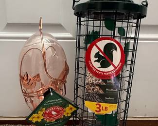 This lot features two bird feeders: a Perky-Pet Looking Glass Hummingbird Feeder and a Perky-Pet Squirrel Stumper 3 lb capacity tube feeder. The hummingbird feeder has a clear glass reservoir with a copper-colored metal base and decorative leaf accents. It is designed to attract hummingbirds and is labeled "Looking Glass Hummingbird Feeder" and "Perky-Pet, Est. 1958, No. 1 in Bird Feeders."
