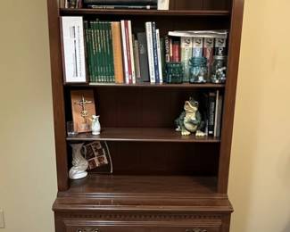 This is a substantial and elegant Ethan Allen French Country tall open bookcase with an integrated set of three drawers. The upper section features adjustable shelves within an arched top cabinet, providing ample display and storage space for books and decorative items. The lower section comprises three drawers, each with decorative brass-toned hardware, offering concealed storage. The overall construction appears to be solid wood with a rich, warm wood finish, likely cherry or mahogany, indicative of Ethan Allen's commitment to quality and traditional design. The piece exhibits classic lines and would complement a variety of interior styles, from traditional to transitional.