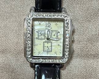 This is a women's fashion watch by Vivani Accutime, featuring a square case adorned with rhinestones. The dial has a mother-of-pearl finish with three subdials for chronograph functions and Arabic numeral '12'. The watch is powered by a quartz movement and has a black patent leather strap. The case back is made of stainless steel and indicates it was made in China with a Japan movement. The overall style is contemporary and decorative, designed for casual wear.