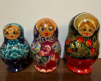 This set features three traditional Russian Matryoshka nesting dolls. The largest doll on the right has a black background with red strawberries and green leaves, and a golden headscarf. The middle doll is painted in shades of purple and red with floral patterns and a blue bow tie. The smallest doll on the left is deep blue with intricate white floral designs and a delicate headscarf. These dolls are typically made of wood, with each subsequent doll nesting inside the previous one. The painting style suggests a folk art tradition, likely originating from Russia.