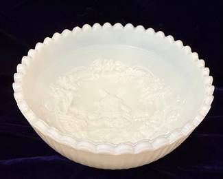 This is a vintage Imperial Glass milk glass bowl featuring the "Windmill" pattern, also known as "Holland" or "Dutch Scene." The pattern depicts a pastoral landscape with a windmill, fence, and floral elements in raised relief. The bowl has a scalloped rim and a milky white, opaque finish characteristic of milk glass. The interior of the bowl is smooth, while the exterior showcases the detailed embossed design. The presence of a maker's mark, appearing to be an "IG" or similar intertwined monogram, is visible on the interior base, consistent with Imperial Glass production.