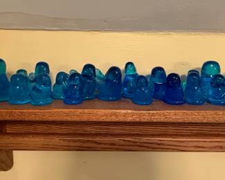 This collection features a group of blue glass bird figurines. They vary in size and shade of blue, from turquoise to deep cobalt. The glass appears solid with a smooth, polished finish. The forms are stylized, representing birds in simplified, elegant silhouettes, with distinct tails and beaks.