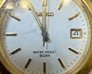 This lot features two men's Seiko wristwatches. The first is a solar-powered watch with a dark blue dial, gold-tone hour markers and hands, and a day-date complication. It indicates 'SOLAR 100M' on the dial and 'MOV'T JAPAN V158-0AD0 R 2' on the case back, which is stainless steel. The second watch has a white dial with gold-tone hour markers and hands, and a day-date complication, and 'WATER RESIST 50M' printed on the dial. Its case back indicates 'MOV'T JAPAN V742-2161 R 2'. Both watches have gold-tone metal bracelets and cases. The overall aesthetic suggests a classic, functional design typical of Seiko's offerings in the late 20th or early 21st century. The crystals on both watches show significant scratching.