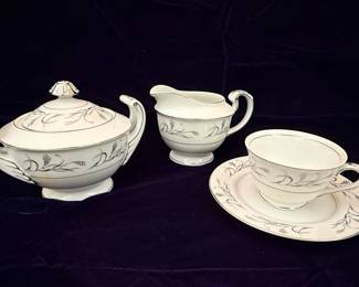 This set includes a lidded sugar bowl, a creamer pitcher, and a teacup with its matching saucer, all crafted from fine china. The pattern, "Platinum Garland," features delicate platinum trim along the edges and a graceful design of wheat stalks and leaves in a muted gray and subtle copper hue. The pieces are white with a smooth, glossy finish, indicative of quality porcelain. The sugar bowl has elegant, curved handles and a decorative lid finial. The creamer exhibits a classic spout and a single handle, while the teacup has a matching handle and sits on a saucer adorned with the same garland motif. The maker's mark on the underside of the teacup identifies it as Harmony House Fine China, Platinum Garland, Made in Japan, with the pattern number 3541.