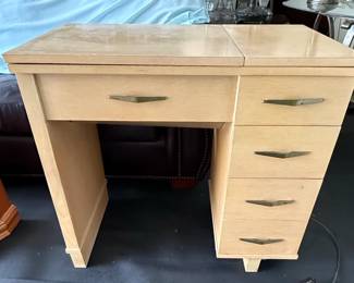 This is a vintage Necchi Supernova sewing machine cabinet, likely from the 1950s or 1960s. It is constructed from light-colored wood, possibly oak or a similar veneer, with a smooth, finished surface. The cabinet features a fold-out top that would conceal a sewing machine, and a set of drawers on the right side for storage. The drawer pulls are a distinctive mid-century modern brass or brass-toned metal. A foot pedal, also with a vintage aesthetic, is visible connected to the unit. The overall design suggests a functional piece of furniture designed to integrate a sewing machine into a living space. A metal plate inside the cabinet reads "ANOTHER NECCHI® CABINET" and includes a serial number.