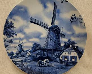 This decorative plate features a classic Dutch landscape scene with windmills, a farmhouse, and grazing cows, rendered in a traditional blue and white Delftware-inspired style. The plate is made of ceramic and is hand-decorated, as indicated by the maker's mark. The design evokes the picturesque scenery of the Netherlands, a common theme in decorative ceramics from the region. The overall aesthetic is rustic yet refined, suitable for display as a collectible or as part of a themed dinnerware set.