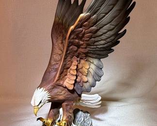 This is a finely crafted porcelain figurine of a bald eagle, a symbol of strength and freedom, captured in mid-descent with wings spread wide. The eagle is perched on a textured rock base, rendered with meticulous detail in its feathers, talons, and piercing gaze. The piece is made of porcelain and features a realistic color palette with browns, whites, and hints of gold on the talons. The underside bears the "Masterpiece Porcelain by HOMCO" mark, "MADE IN MEXICO," and the copyright year "1986."
