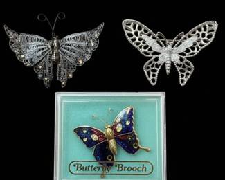 This lot features three distinct vintage butterfly brooches. The largest, presented in its original 'Butterfly Brooch' box, is a colorful enamel and rhinestone piece with iridescent blue wings accented with red and yellow, and small clear rhinestones against a gold-toned body. The other two brooches are silver-toned metal. One is a delicate filigree piece with intricate, lace-like detailing on its wings, adorned with tiny clear rhinestones. The third is an open-work filigree design with a textured body and wings featuring a honeycomb-like pattern. All three appear to be costume jewelry, likely dating from the mid to late 20th century.