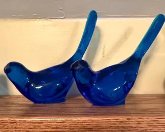 This collection features a group of blue glass bird figurines. They vary in size and shade of blue, from turquoise to deep cobalt. The glass appears solid with a smooth, polished finish. The forms are stylized, representing birds in simplified, elegant silhouettes, with distinct tails and beaks.