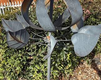This is a metal kinetic wind sculpture, designed for garden or outdoor display. It features stylized tulip petal forms that rotate with the wind, along with stylized leaf forms. The material appears to be weathered steel or a similar metal, exhibiting a patina and some signs of rust, which is common for outdoor metal art exposed to the elements. The sculpture is mounted on a central pole, suggesting it is intended to be anchored in the ground.