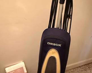 This is an Oreck XL Gold upright vacuum cleaner, a model known for its lightweight design and powerful suction. The vacuum features a durable construction with a dark blue fabric bag, accented with gold and black trim. The 'XL GOLD' designation is prominently displayed on the unit, indicating a specific series within Oreck's product line. It is designed for efficient cleaning of carpets and bare floors. The accompanying filter bag appears to be a standard replacement part for Oreck vacuums.