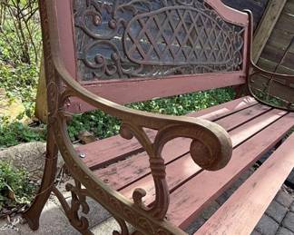 This garden bench features a decorative cast iron frame with scrolled armrests and a central back panel adorned with an intricate, lattice-like pattern. The seat and backrest are constructed from wooden slats, painted a muted reddish-brown. The cast iron elements exhibit a weathered, aged patina, suggesting outdoor use. The overall design evokes a Victorian or early 20th-century aesthetic, suitable for garden or patio seating. This specific bench appears to be the 'Tea Rose' model by Oakland Living.