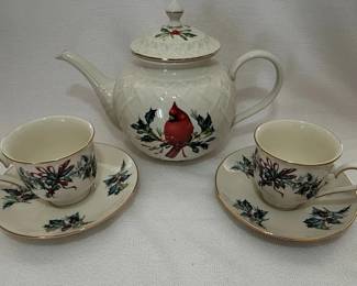 This Lenox Winter Greetings set features a white Fine Ivory China teapot and two cups, each adorned with a cardinal perched amidst holly and berries. The teapot has a distinctive quilted pattern on its body and a decorative finial. The cups share the same festive motif and are accented with a gold rim. The set is made of fine ivory china, known for its durability and translucent quality. The design evokes a classic holiday or winter theme. The maker's mark on the cups indicates 'Lenox Fine Ivory China, Made in U.S.A., 1995'.