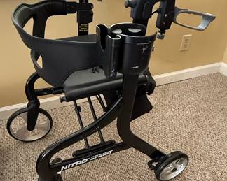 This is a Drive Nitro "Sprint" rollator walker, designed for mobility assistance. It features a lightweight aluminum frame in a black finish, with a padded seat and backrest for comfort. The rollator has large 6-inch front and rear casters for smooth maneuverability over various surfaces. It includes adjustable ergonomic handles, a removable storage pouch, and a cup holder. The "Sprint" model is known for its compact and lightweight design, making it easy to fold for transport and storage.