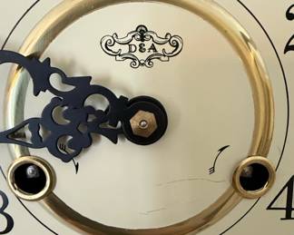 This is a wooden mantel clock featuring a "wheat sheaf" motif. The clock face is cream-colored with black Arabic numerals and decorative black hands. A gold-toned bezel surrounds the glass cover. The maker's mark "D&A" is visible on the clock face, and a "Made in China" sticker is present on the underside, along with a model number "9409WPS". The case is constructed from wood with a light to medium oak finish, exhibiting a traditional arched top and a stepped base. The decorative elements suggest a classic or rustic aesthetic.