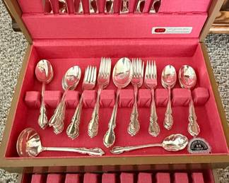 This is a vintage 1847 Rogers Bros silverplate flatware set in the "Reflection" pattern, presented in its original wooden chest lined with pink felt. The "Reflection" pattern, introduced by International Silver Company, is characterized by its elegant, ornate scrollwork and floral motifs on the handle, a style popular in the mid-20th century. The set includes a variety of serving pieces and place setting utensils, indicative of a comprehensive collection designed for entertaining. The silverplate is likely a nickel silver base with a heavy layer of silver electroplated onto it. The chest itself is designed to protect and organize the pieces, featuring individual slots for each utensil.