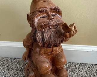 This is a hand-carved wooden figurine depicting a gnome. The gnome is shown in a seated position with its eyes closed, holding a small, four-leaf clover or flower in its outstretched hand. The carving exhibits a rustic style with detailed features such as a long beard, textured clothing, and a pointed hat. The wood has a warm, natural finish with visible grain patterns. The base appears to be carved to resemble a tree stump or rock. A maker's mark, 'R.E.B. 2-10', is visible on the underside, suggesting it was created by an artist named R.E.B. in February 2010.