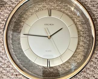 This is a modern Verichron quartz wall clock featuring a minimalist design. The clock has a clean white dial with subtle silver-tone accents and Roman numerals for the hours, except for the 12 and 6 o'clock positions which are marked with simple black lines. The hands are slender and dark, with a gold-tone central hub. The clock is encased in a clear acrylic or glass dome, which is then surrounded by a polished silver-tone metal bezel. The overall aesthetic is sleek and contemporary, suitable for various interior decors.