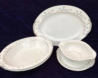 This set includes a lidded sugar bowl, a creamer pitcher, and a teacup with its matching saucer, all crafted from fine china. The pattern, "Platinum Garland," features delicate platinum trim along the edges and a graceful design of wheat stalks and leaves in a muted gray and subtle copper hue. The pieces are white with a smooth, glossy finish, indicative of quality porcelain. The sugar bowl has elegant, curved handles and a decorative lid finial. The creamer exhibits a classic spout and a single handle, while the teacup has a matching handle and sits on a saucer adorned with the same garland motif. The maker's mark on the underside of the teacup identifies it as Harmony House Fine China, Platinum Garland, Made in Japan, with the pattern number 3541.