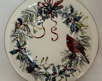 This Lenox plate, part of the Millennium Edition, features the 'Winter Greetings' design by artist Catherine McClung. The plate is crafted from fine ivory china and made in the U.S.A. The front of the plate depicts a festive winter scene with a wreath of pine, holly, and mistletoe, adorned with a red ribbon and featuring a cardinal and a chickadee. The rim is accented with a delicate gold band. The back of the plate displays the artist's signature, the Lenox logo, and details about the edition and year of production, 1995. This is specifically a dinner plate, as indicated by the size and the label on the back.