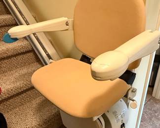 This is a Handicare Simplicity Seat RH stairlift, designed to provide safe and comfortable mobility assistance on straight staircases. The seat is upholstered in a neutral beige fabric, featuring padded armrests that fold up for easy access. A safety belt is integrated for user security. The control unit, which includes up (green) and down (red) buttons, is operated via a key for security and can be mounted on either side of the chair. The unit is powered by a rechargeable battery system, ensuring operation during power outages. The 'RH' designation likely indicates 'Right Hand' side installation relative to ascending the stairs.