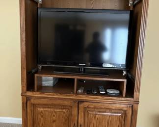 This large entertainment center armoire is constructed from solid oak, featuring a warm, medium-toned finish that highlights the natural grain of the wood. The design incorporates a central open section designed to house a television, with a smaller shelf below for media components. The lower section features two paneled doors with traditional hardware, opening to reveal shelved storage. The overall style is transitional, blending elements of traditional craftsmanship with a functional design suitable for modern media setups. The piece exhibits sturdy construction with visible joinery, suggesting it is a well-made, durable furniture item.