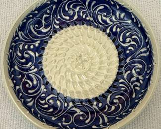 This is a decorative and functional ceramic plate designed for grating garlic. It features a central textured surface with raised ridges, perfect for mincing garlic cloves by rubbing them against the abrasive center. The rim is adorned with a traditional blue and white swirling foliate pattern, characteristic of Spanish ceramics. The underside of the plate bears a hand-written inscription, 'Triana Sevilla,' indicating its origin from the Triana district of Seville, Spain, a region renowned for its pottery production. The plate is likely made from earthenware or stoneware, finished with a glossy glaze.