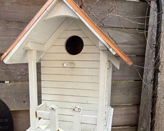 This charming white wooden birdhouse features a classic pitched roof design with a gleaming copper accent, reminiscent of the Cape Cod Wren style. The house is constructed from painted wood, with horizontal siding on the main body and supporting posts for a small porch area. A circular entrance hole is centrally placed on the front facade, and a perch extends below it. The copper roof shows signs of patina, indicating exposure to the elements. The overall construction suggests a functional and decorative garden ornament, likely intended for small backyard birds.