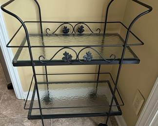 A vintage two-tiered bar cart made of black wrought iron, featuring decorative leaf motifs and scrollwork. The shelves are textured glass. The cart has curved handles and is supported by four legs, two with casters.