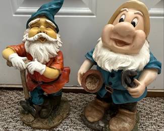 This set features two garden statues: one depicting Grumpy from Disney's Snow White and the Seven Dwarfs, and the other a classic gnome figure. Both are crafted from plaster and hand-painted. Grumpy is shown in his signature blue tunic and red cap, holding a small brown hat and a horseshoe. The gnome, with a long white beard and a pointed blue hat, leans on a wooden staff that resembles a shovel. They are designed to add a whimsical touch to any garden or outdoor space.